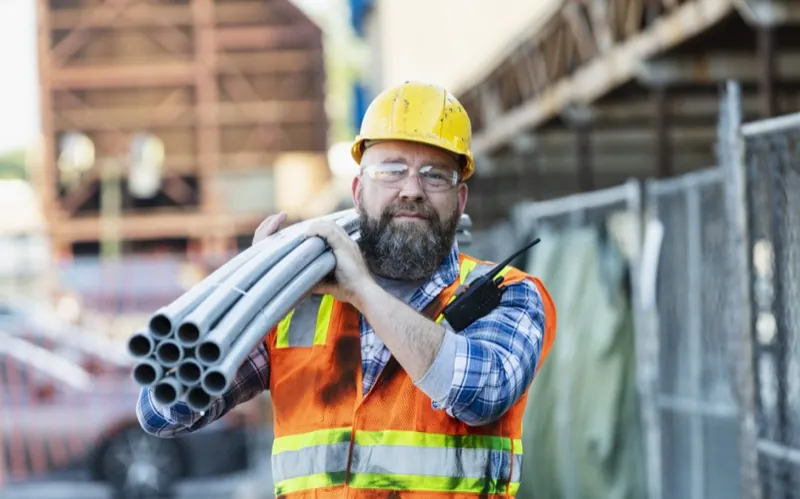 Construction worker carrying pipes on an active jobsite wearing hi-vis vest and hard hat — construction time tracking